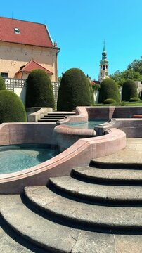 Prague Loreta. Serene garden landscape featuring elegant topiary hedges, stone steps leading to a tranquil water feature, and a historic tower in the background under a clear blue sky