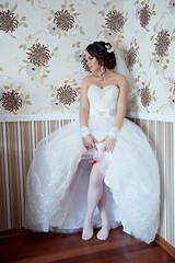 A bride is wearing a white dress and red stockings. She is standing in front of a wall with floral...