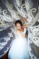 A woman in a white dress is posing in front of a lace curtain. The curtain is white and has a...