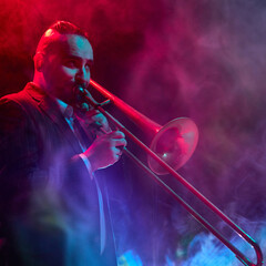 A musician, Caucasian descent, plays the trombone under vibrant red and blue stage lights with...