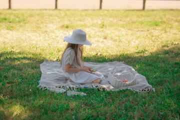 A young girl in a wide-brimmed hat sits peacefully on a picnic blanket with a basket, embodying the...