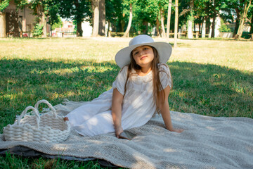 A young girl in a wide-brimmed hat sits peacefully on a picnic blanket with a basket, embodying the...
