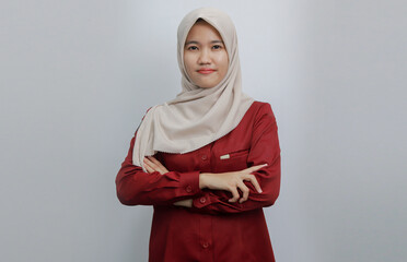 A confident Muslim woman wearing a beige hijab and red shirt poses with her arms crossed against a...