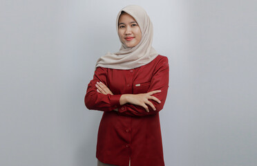 Fototapeta premium A confident Muslim woman wearing a beige hijab and red shirt poses with her arms crossed against a plain background.