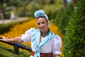 Portrait of young, beautiful, blonde woman, with typical Andalusian flamenco costume to go on pilgrimage in white color with light blue shawl and flower, leaning on a railing in an outdoor park.