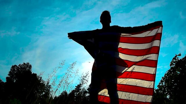 A person as a silhouette holding up an American United States flag in nature while wind is moving it slowly and smoothly wrapping around the person. Blue sky and nature is visible in the background.