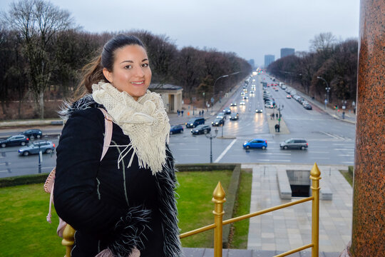 Young woman in winter outfit smiling at the camera, standing at the Victory Column in Berlin Germany with a busy street, cars and leafless trees in the background.
