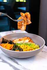 Fork holding grilled chicken above poke bowl with avocado and vegetables