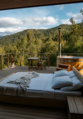 An outdoor deck with a bed, pillows, light blanket, a rustic table and chair, and a wooden hot tub overlooking lush forested mountains under a blue sky with scattered clouds. 