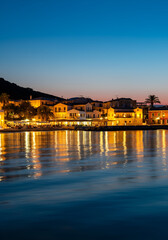 A seaside village with illuminated Mediterranean-style houses reflecting on calm evening waters, captured during blue hour with a gradient sky fading from sunset.