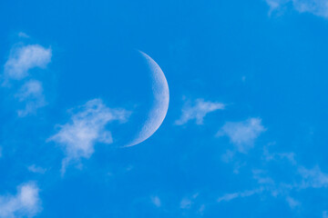 A delicate crescent moon shines in the clear blue sky, surrounded by soft white clouds. A peaceful daytime celestial view with crisp atmospheric contrast.