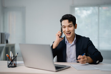 Young, happy businessman talking on mobile phone while working on laptop, smiling at the screen, holding a pen, and multitasking in a modern office