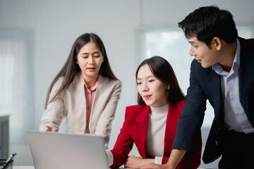 Three asian businesspeople are discussing a project while looking at a laptop in a modern office,...