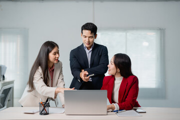 Three asian businesspeople are discussing work, using a laptop in a modern office, collaborating on...