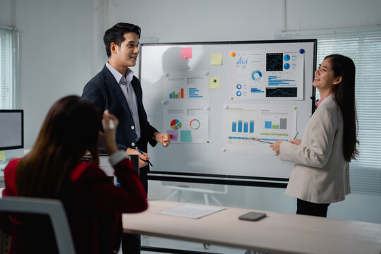 Two asian businesspeople discussing company strategy during a meeting in a modern office, using a whiteboard and financial reports, while a colleague is listening