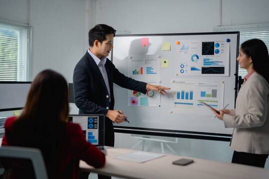 Asian businessman in suit pointing at whiteboard presenting company strategy and showing marketing research results to colleagues in modern office