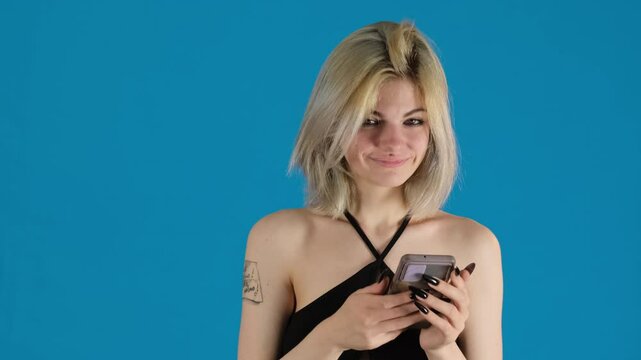 Young woman brightly smiles at phone on blue background. Lady in black top calls out for someone to come and watch eager to share funny message