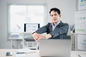 Fototapeta premium Asian businessman in suit stretching arms at workplace with laptop on desk, working on computer in office, tired manager taking break from work, business concept
