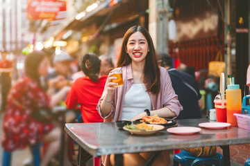 Smiling face traveler woman cheers beer eating pad thai at street food market foodie asia tour in Chinatown Bangkok city