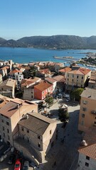 Vue sur la place de la R&eacute;publique &agrave; Porto-Vecchio, entour&eacute;e de maisons en pierre et de b&acirc;timents color&eacute;s, avec les tables d&rsquo;un restaurant install&eacute;es en terrasse. En arri&egrave;re-plan, le port de plaisance 