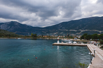 Fototapeta premium Scenic view of the harbor of Ancient Epidaurus in Greece, with lighthouse, fishing boats, pier, waterfront walkway, and mountains in the background under cloudy sky.