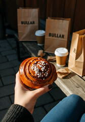 A slightly tilted top-down photo of a hand holding a Swedish cinnamon bun topped with pearl sugar. Set in a rustic outdoor caf&eacute; environment with a wooden bench, kraft paper bags, and takeaway coffee.