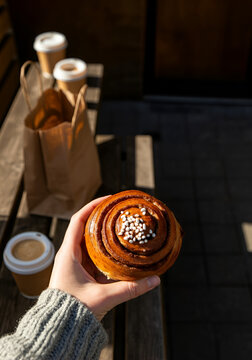 Slightly tilted top-down photo of a hand holding a Swedish cinnamon bun topped with pearl sugar. Set in a rustic outdoor caf&eacute; environment with a wooden bench.