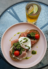 An open-faced toast with prosciutto, poached egg, cherry tomatoes, olives, and a green herb sauce served on a pastel pink plate, accompanied by iced tea with lime slices on a glass tabletop.