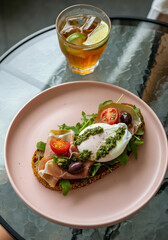 An open-faced toast with prosciutto, poached egg, cherry tomatoes, olives, and a green herb sauce served on a pastel pink plate, accompanied by iced tea with lime slices on a glass tabletop.