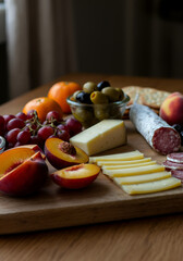 A rustic charcuterie board with sliced cheese, fruit crackers, tangerines, fresh plums, peaches, salami, and a glass dish of green and black olives, set on a wooden surface. 