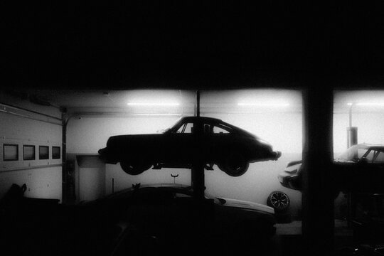 Front view of a classic black Porsche 911 Carrera parked in a dimly lit vintage car workshop next to a yellow sports car, capturing the nostalgic atmosphere of a restoration garage.