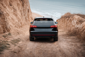 Black SUV driving down a hill to a beach. Sporty SUV on a dirt road near sandy cliffs by the sea. 