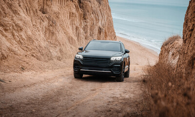 Black SUV driving uphill from a beach near a cliff. Sporty SUV on a dirt road with sandy soil near...