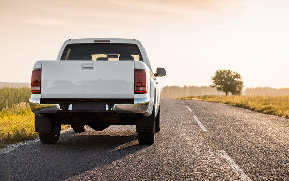 White pickup truck on a countryside paved road in late afternoon. Rear view of white truck against the sun, lone tree on the right. 