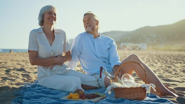 Retired couple sharing intimate moment while pouring white wine, sitting on beach blanket with picnic basket, enjoying fresh fruits during golden sunset by ocean shore