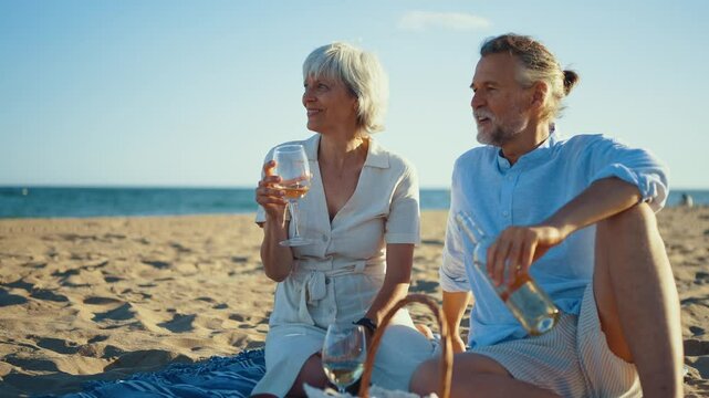 Retired couple sharing intimate moment while pouring white wine, sitting on beach blanket with picnic basket, enjoying fresh fruits during golden sunset by ocean shore
