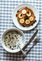 Top-down view of a healthy breakfast scene featuring a bowl of milk with sunflower seeds, pumpkin seeds, and raisins, paired with a plate of pancakes topped with sliced bananas and raisins. 