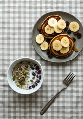 Top-down view of a healthy breakfast scene featuring a bowl of milk with sunflower seeds, pumpkin seeds, and raisins, paired with a plate of pancakes topped with sliced bananas and raisins. 