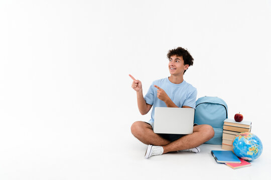 Fototapeta Amazed boy guy high school male pupil student teenager sitting near books copybooks backpack globe using laptop computer for e-learning pointing index fingers copy space isolated on white background.