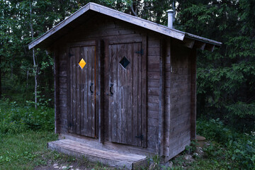 An old, rustic double outhouse made of dark wood in a dense forest at twilight. A light is on in one of the cabins. A secluded latrine.