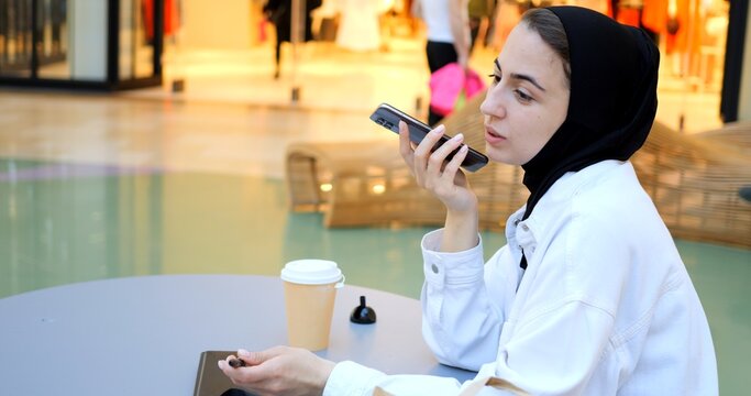 Young muslim businesswoman wearing hijab sitting at table in shopping mall, recording voice message on smartphone, holding pen and takeaway coffee cup next to tablet. Free and modern Muslim in city