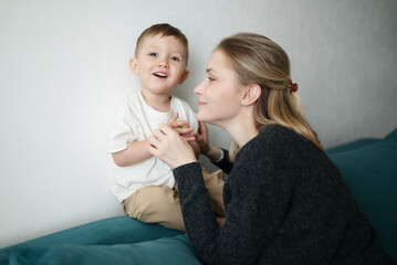 Cheerful young mother and her little son smiling and bonding while sitting together on a teal couch...