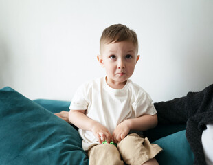 Cute little boy with a funny pout sitting on a teal couch at home, wearing a white t-shirt and...