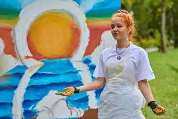 Young female with red hair discusses vibrant ocean sunset painting outdoors. Wearing white apron,...