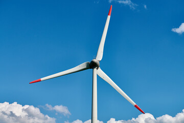 Modern wind turbine with red tipped blades stands tall against vivid blue sky filled with scattered...
