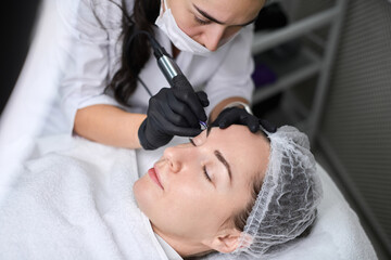 Young woman receives eyebrow permanent makeup in serene spa. Beauty technician, wearing gloves,...