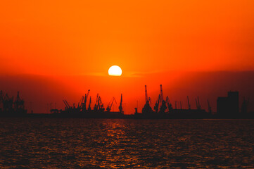 Industrial port cranes silhouetted against vivid orange sunset sky over calm sea, with glowing...
