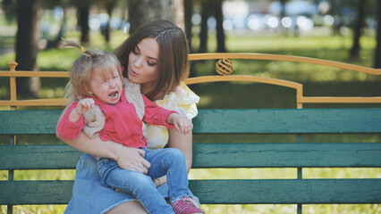 Nurturing mother soothing crying infant while seated on park bench during bright summer afternoon