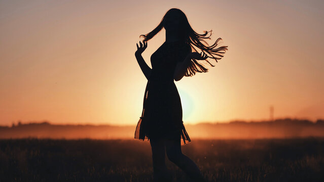 Silhouette of carefree girl dancing in field at sunset, hair flowing in wind, enjoying moment of freedom and happiness