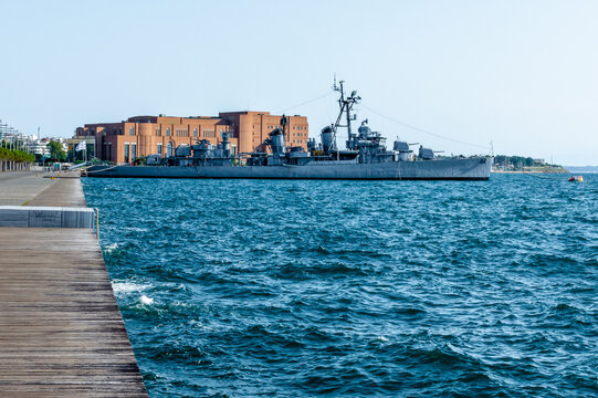 Averof battleship museum docked on the Thessaloniki seafront in Greece, with blue sea, summer light and the modern red concert hall in the background.
